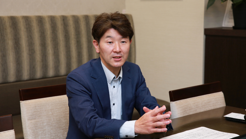 Man in a blue blazer sits at a conference table, hands clasped, speaking in a meeting room with papers in front of him