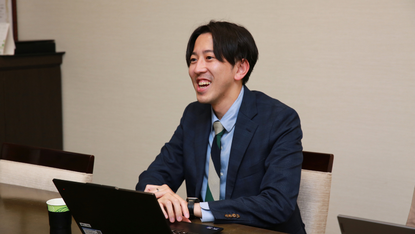 Smiling man in a navy suit and striped tie sits at a meeting table with an open laptop in front of him.