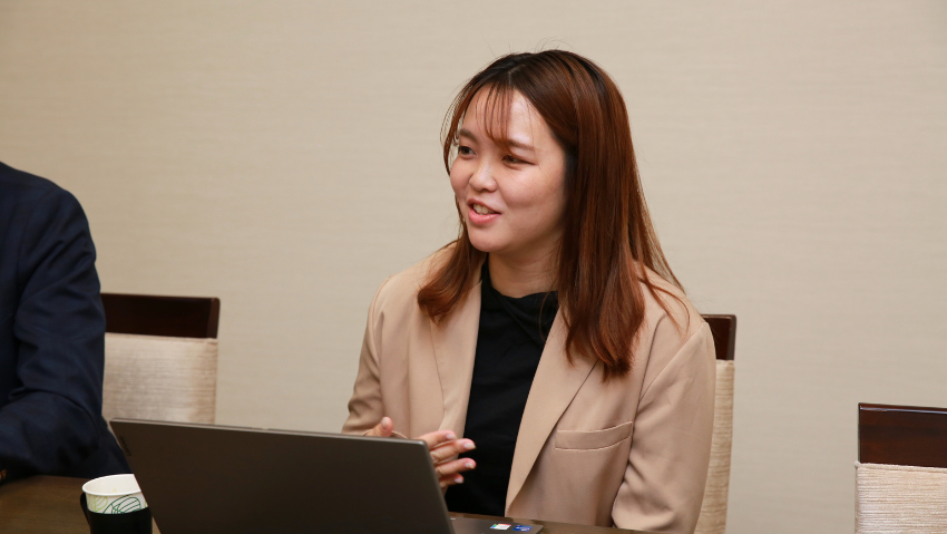 Woman in a beige blazer sits at a table with an open laptop, speaking during a meeting.