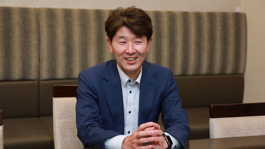 Man in a blue suit sits at a table, smiling with hands clasped in front of him on the table.