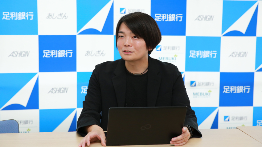 Person in a black blazer sits at a table with a laptop, speaking in front of a blue and white logo backdrop.