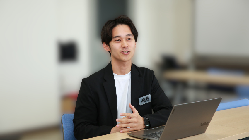 Young man in a black blazer speaks while seated at a desk with a laptop in a classroom setting.