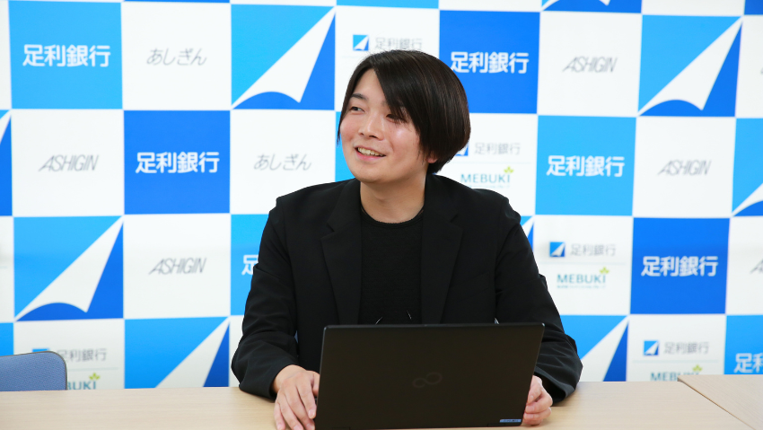 Person in a black blazer sits at a desk with a laptop, smiling, in front of a blue-and-white logo backdrop.