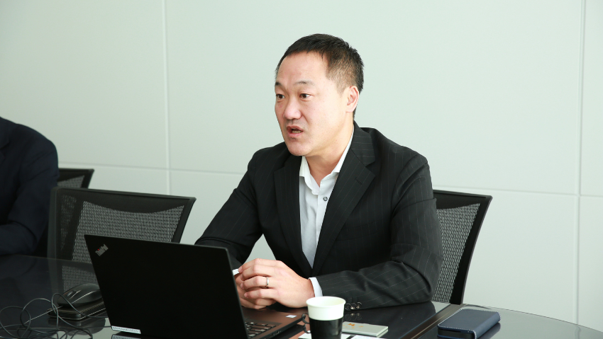 Man in a dark suit speaks while seated at a conference table with a laptop in front of him and a coffee cup nearby.