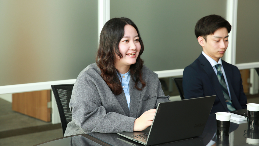 Two professionals sit at a conference table; a smiling woman types on a laptop while a man in a suit looks on nearby.