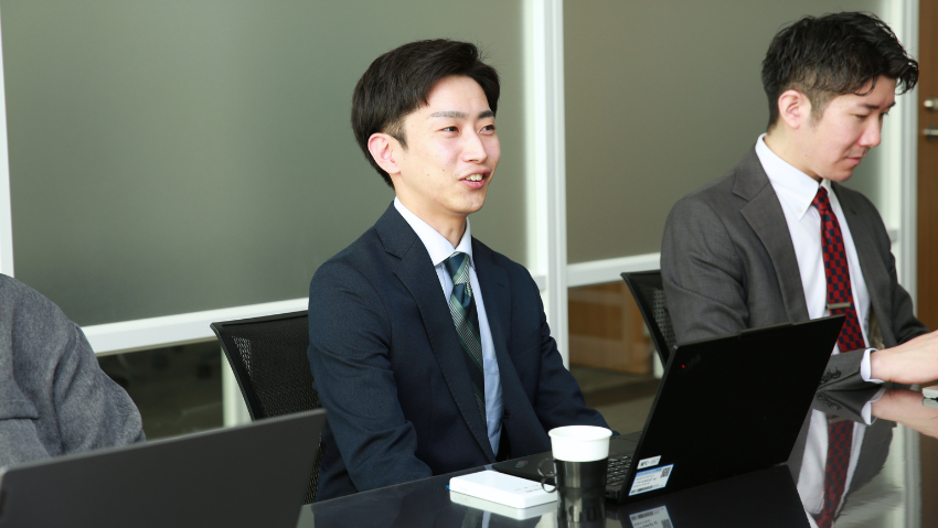 Two businessmen sit at a conference table with laptops; one man in a navy suit speaks while the other adjusts a device nearby.