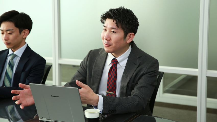 Two businessmen in a conference room: the man in the foreground in a gray suit gestures with his hands while a laptop sits on the table and a coffee cup nearby.