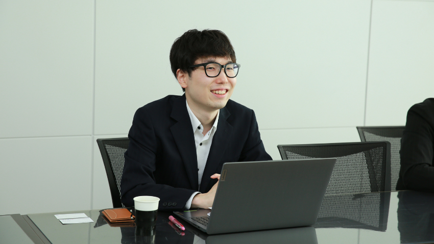 Young man in a suit and glasses sits at a conference table with a laptop, smiling during a meeting.