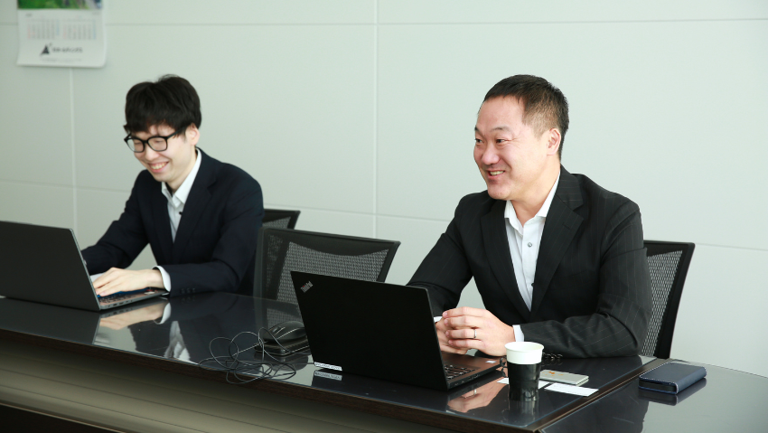 Two businessmen in suits sit at a conference table with laptops, smiling during a meeting over coffee and notes.