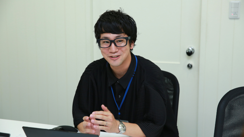 Asian man with short black hair and glasses sits at a desk, hands clasped, smiling in an office setting