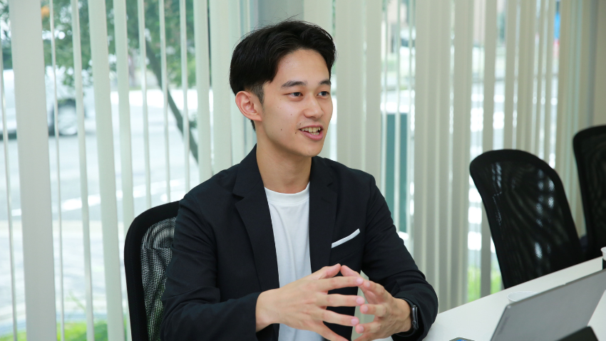 Young man in a dark blazer sits at a conference table and speaks, hands clasped, with a laptop nearby.