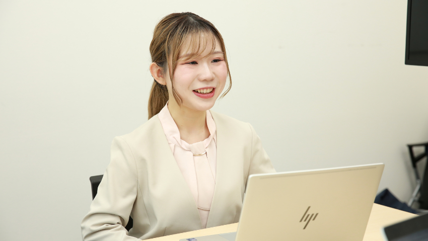 Smiling woman in a beige blazer sits at a desk, working on a silver HP laptop in a neutral office.