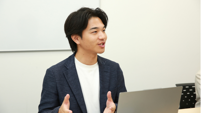Young man in a navy blazer speaks and gesticulates, seated with a laptop in front of him.